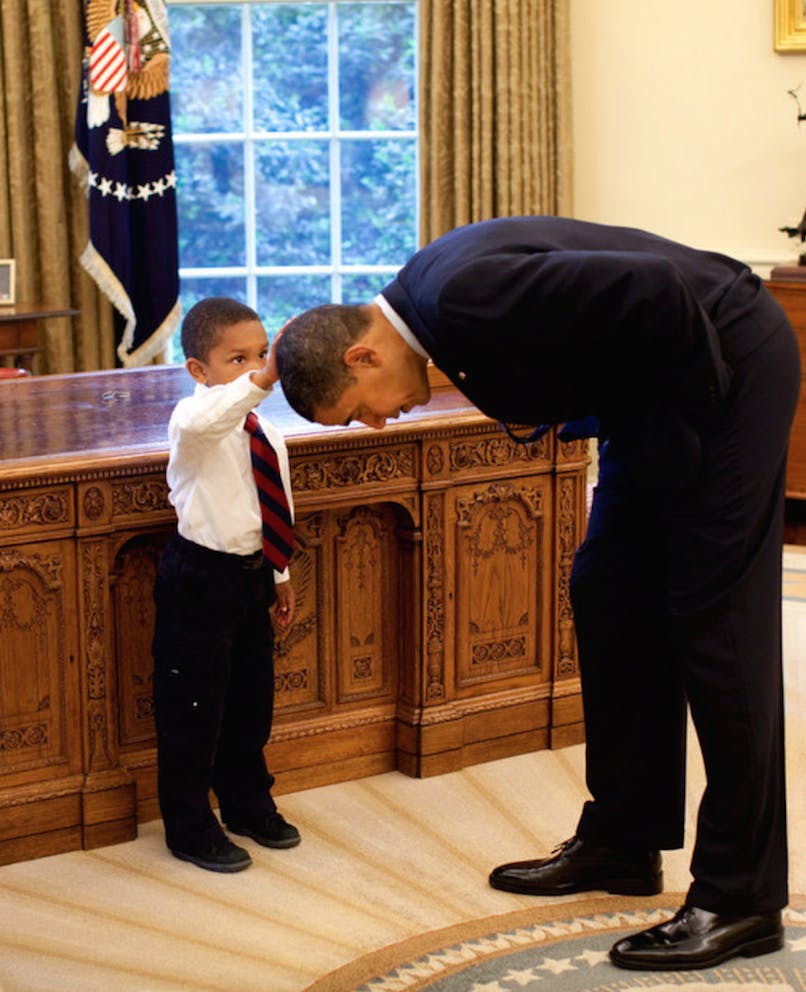 President Barack Obama bends over so the son of a White House staff member can pat his head during a family visit to the Oval Office